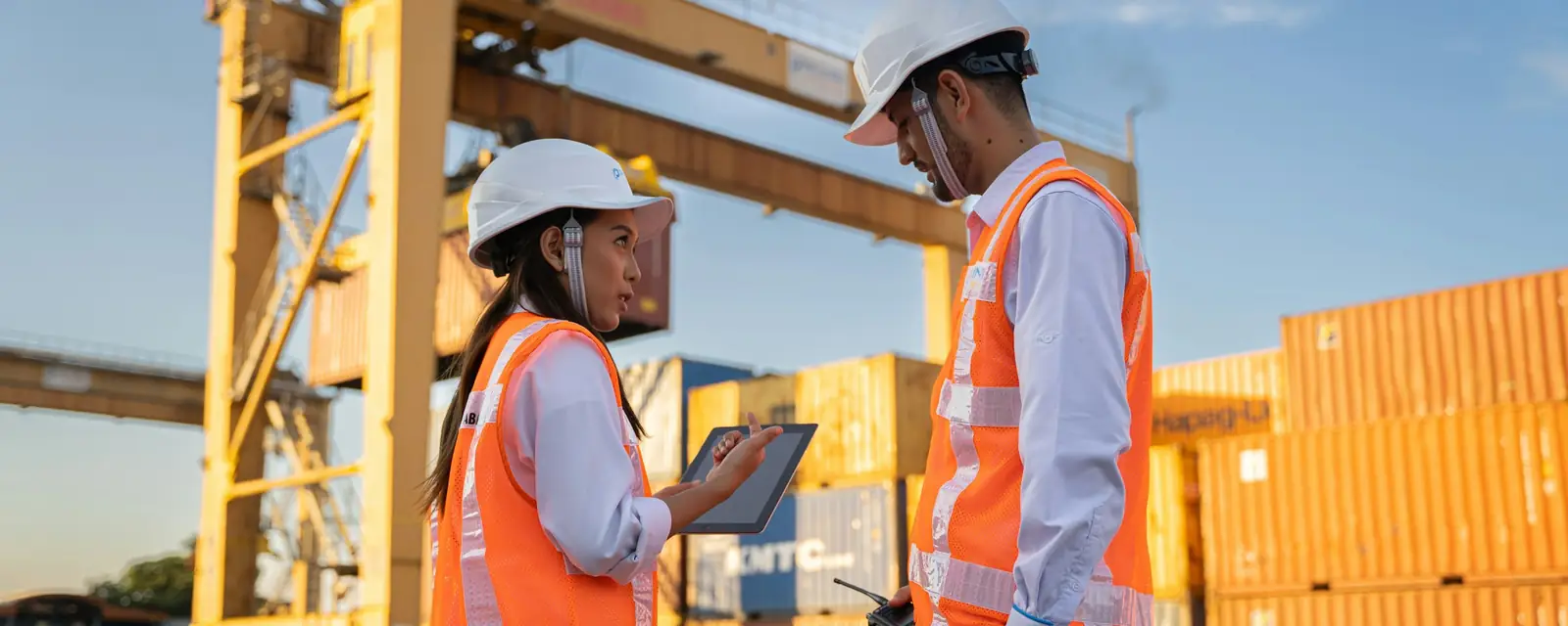 Two logistics workers in hard hats and hi-vis vests checking a tablet beside a gantry crane and stacked containers