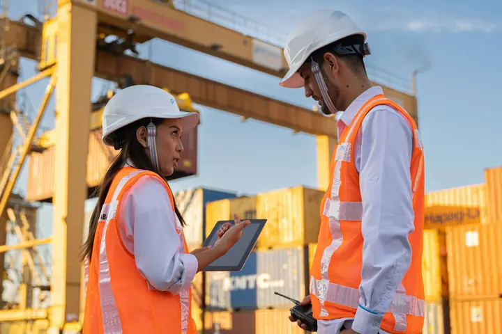 Two logistics workers in hard hats and hi-vis vests checking a tablet beside a gantry crane and stacked containers