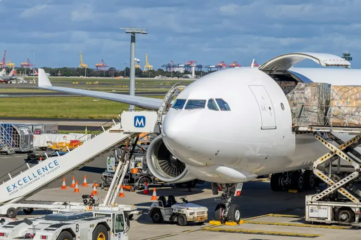 Wide-body freighter aircraft on the apron with its main-deck cargo door open and a pallet being raised on a high loader