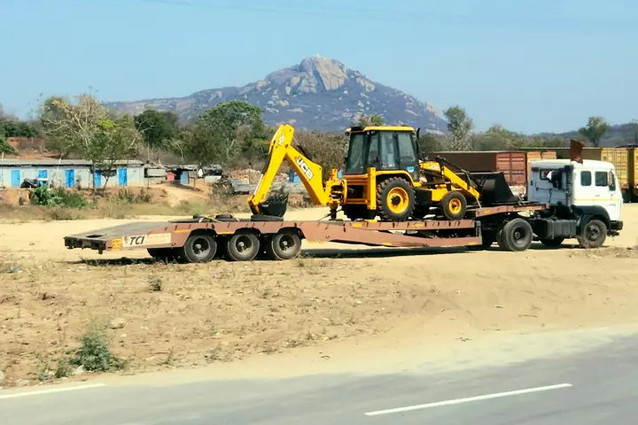 A truck and low trailer carrying a yellow backhoe loader past a building site