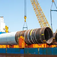 Crawler cranes lifting large steel monopile sections onto the deck of a heavy-lift ship in port