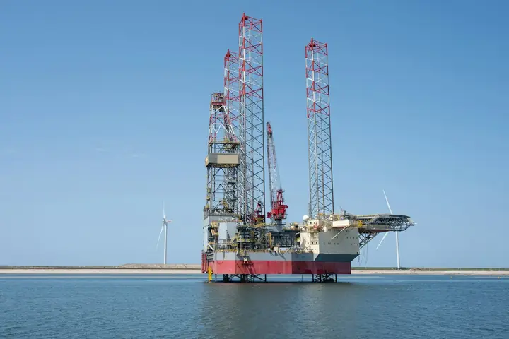 A jack-up drilling rig standing on its legs in calm water, with wind turbines on the horizon