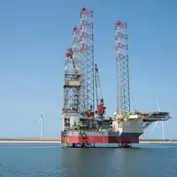 A jack-up drilling rig standing on its legs in calm water, with wind turbines on the horizon