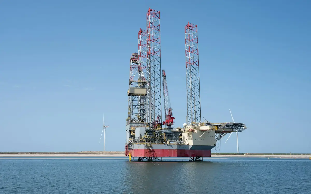 A jack-up drilling rig standing on its legs in calm water, with wind turbines on the horizon