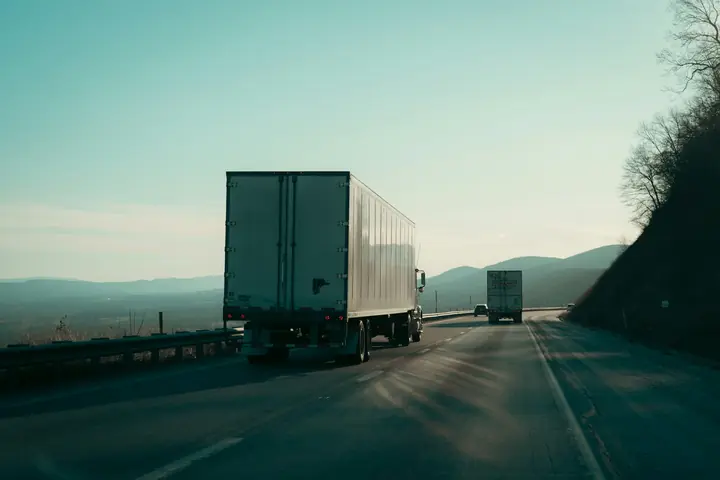 Two articulated box trailers driving along a motorway towards the hills at sunrise